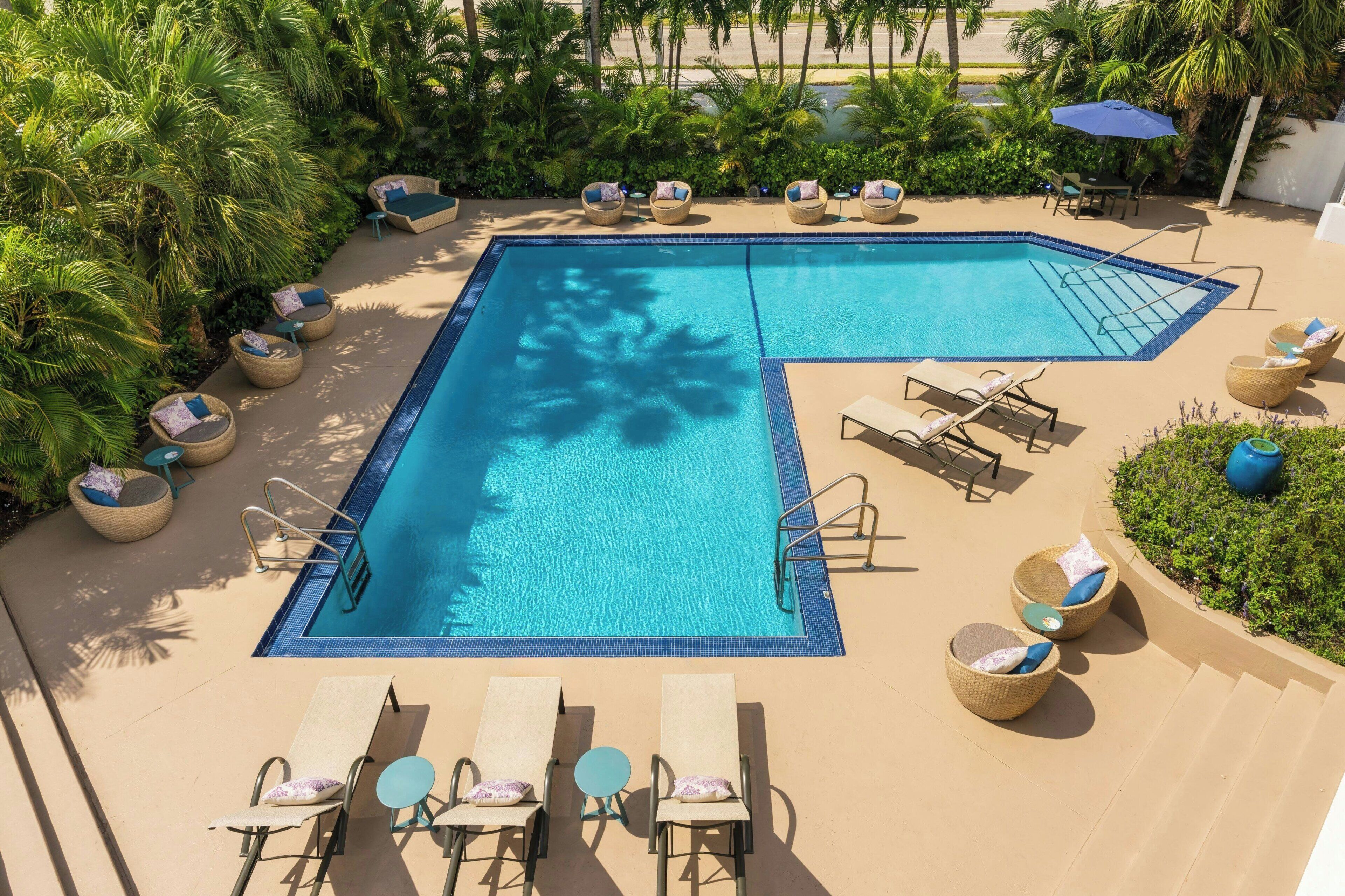 Piscina com formato geométrico, cercada por espreguiçadeiras e mesas, e com palmeiras para dar sombra.