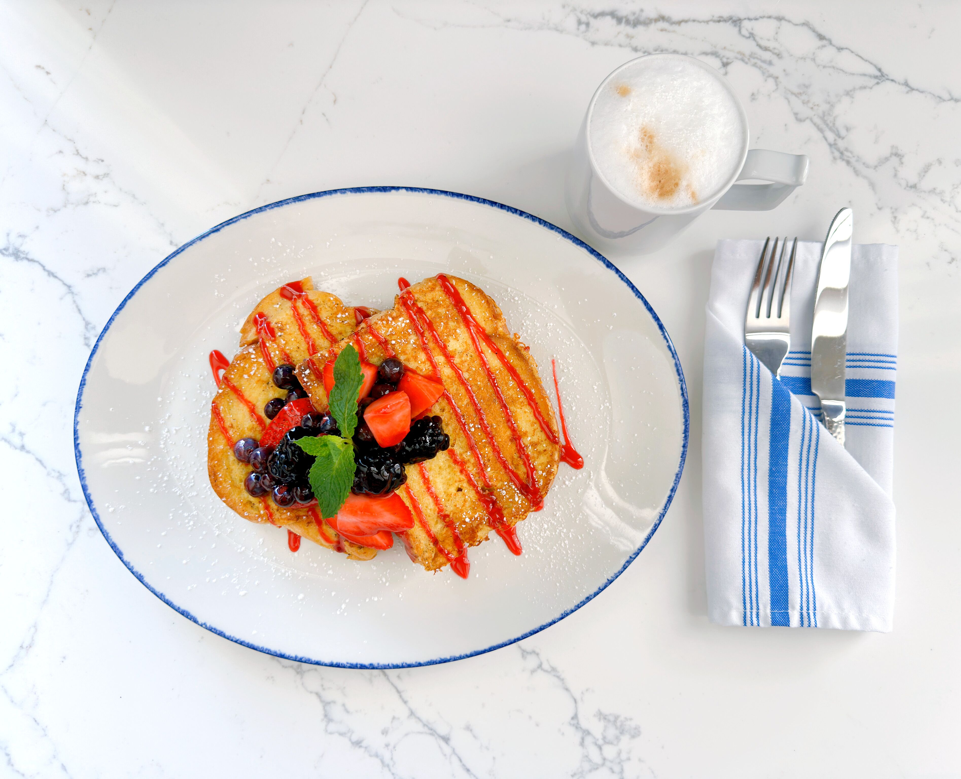 A plate of food with a blue rim, a glass of a beverage, and a striped napkin with cutlery on a marble surface.