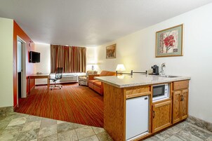 Una habitación de hotel compacta con kitchenette, una zona de estar con sofá y sillón, un televisor y un cuadro en la pared.