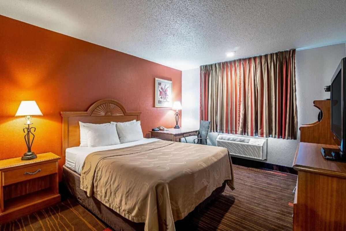A hotel room with a bed, bedside lamps, a desk, a chair, a television, and a window with curtains.