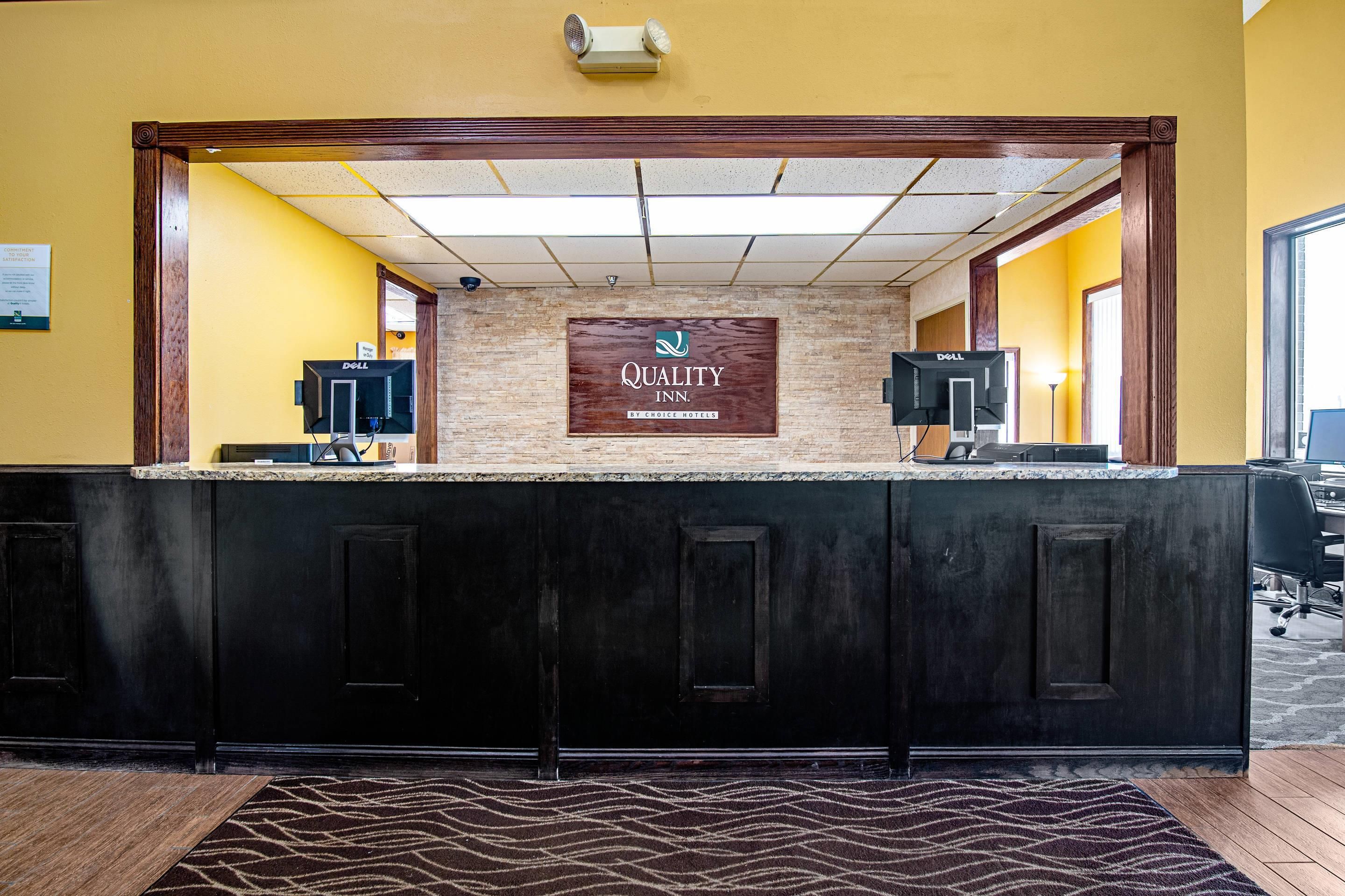 A hotel reception desk with a sign reading 'Quality Inn by Choice Hotels,' two computer monitors, and a patterned rug on the floor.