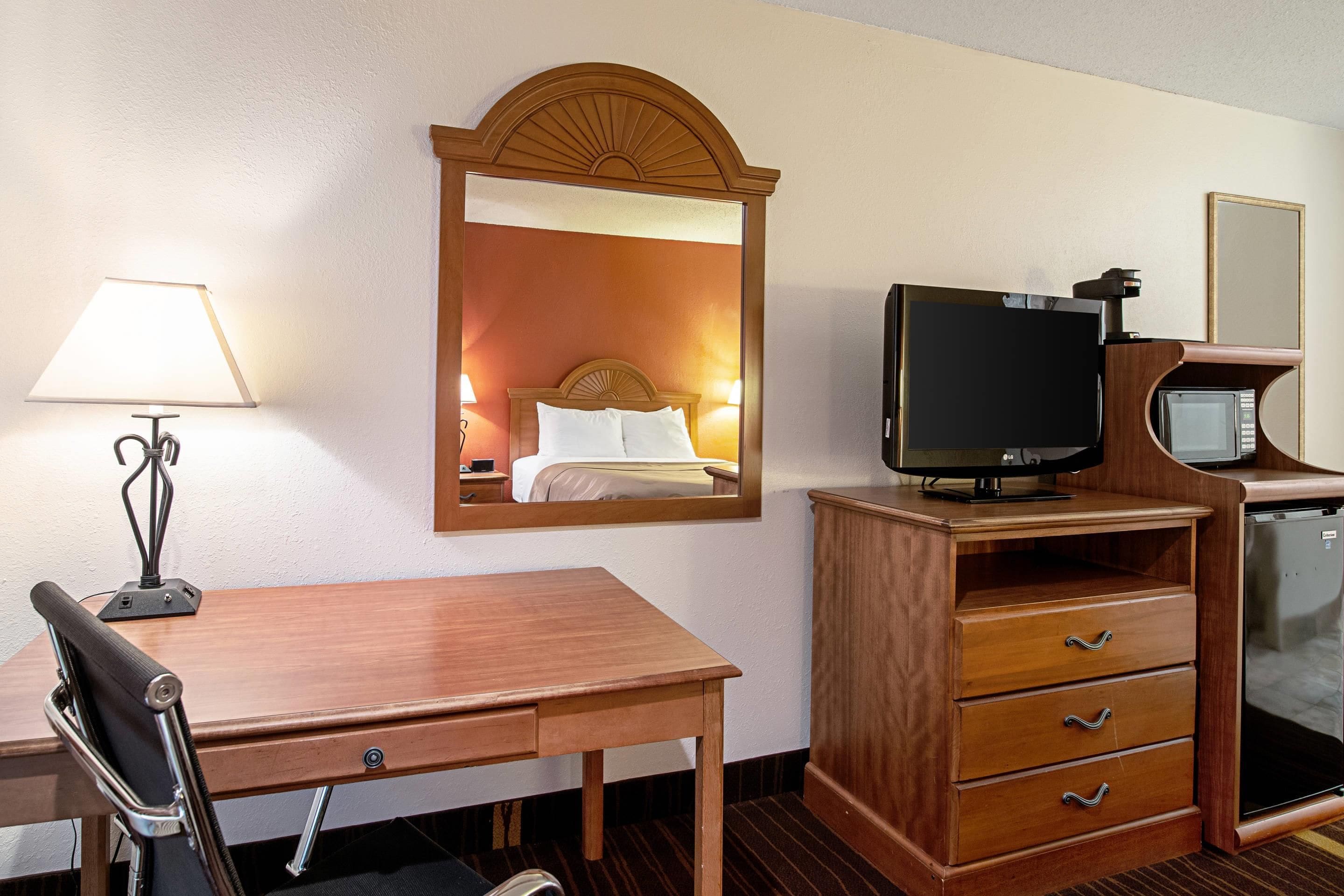 A hotel room with a wooden desk, a chair, a television, a mirror, a lamp, and a minibar.
