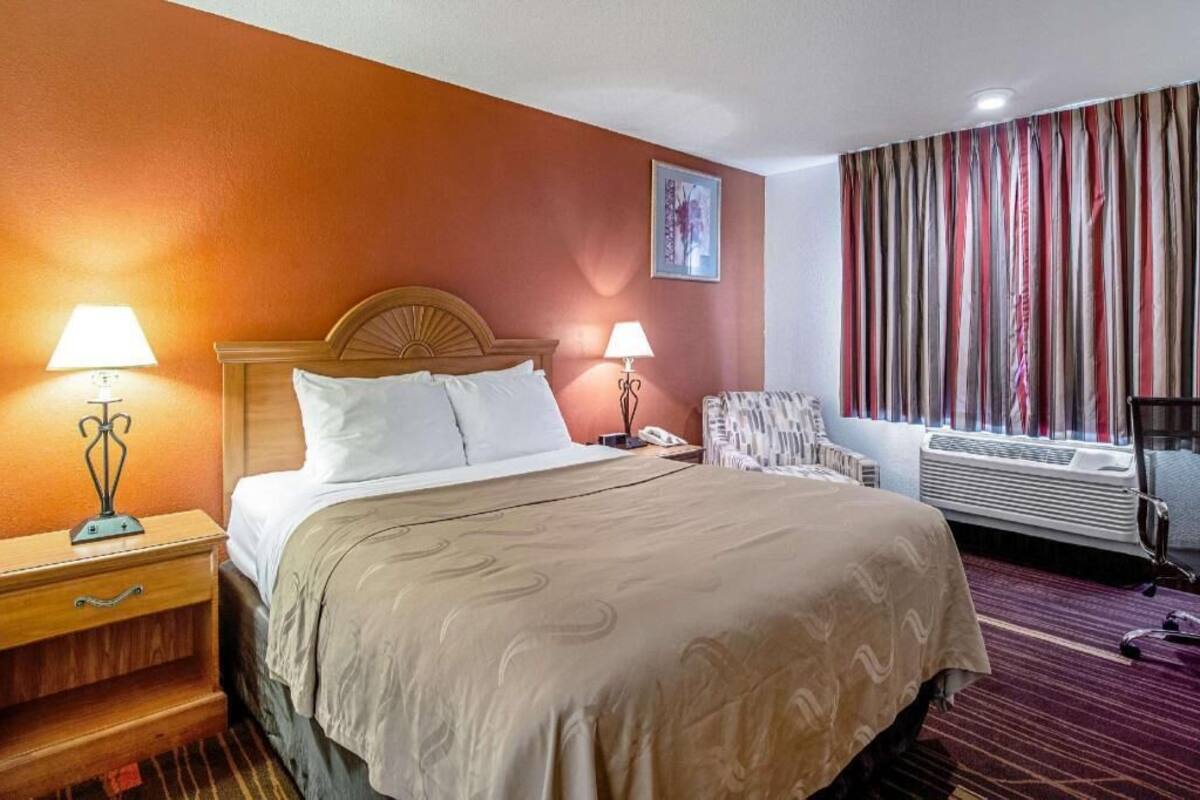A hotel room with a bed, two bedside tables with lamps, a chair, a window with curtains, and an air conditioning unit.
