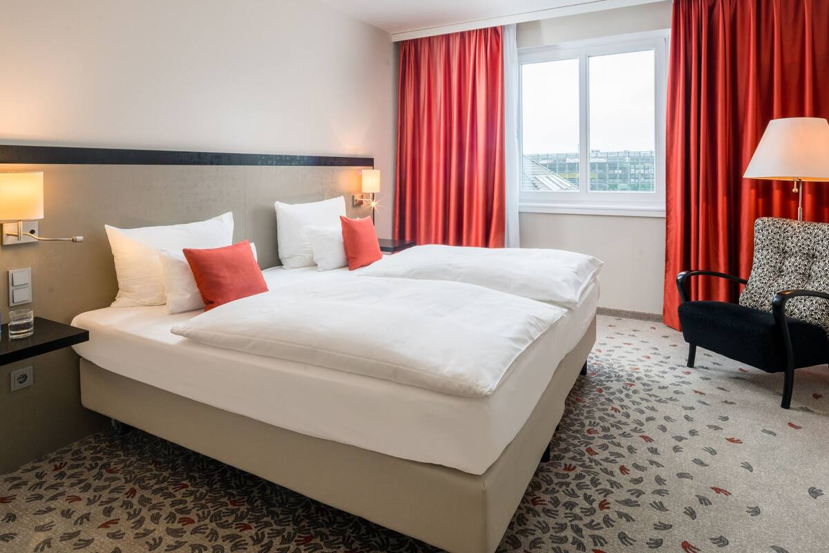 Een hotelkamer met een groot bed, rode gordijnen, een kamerbreed tapijt en een stoel.