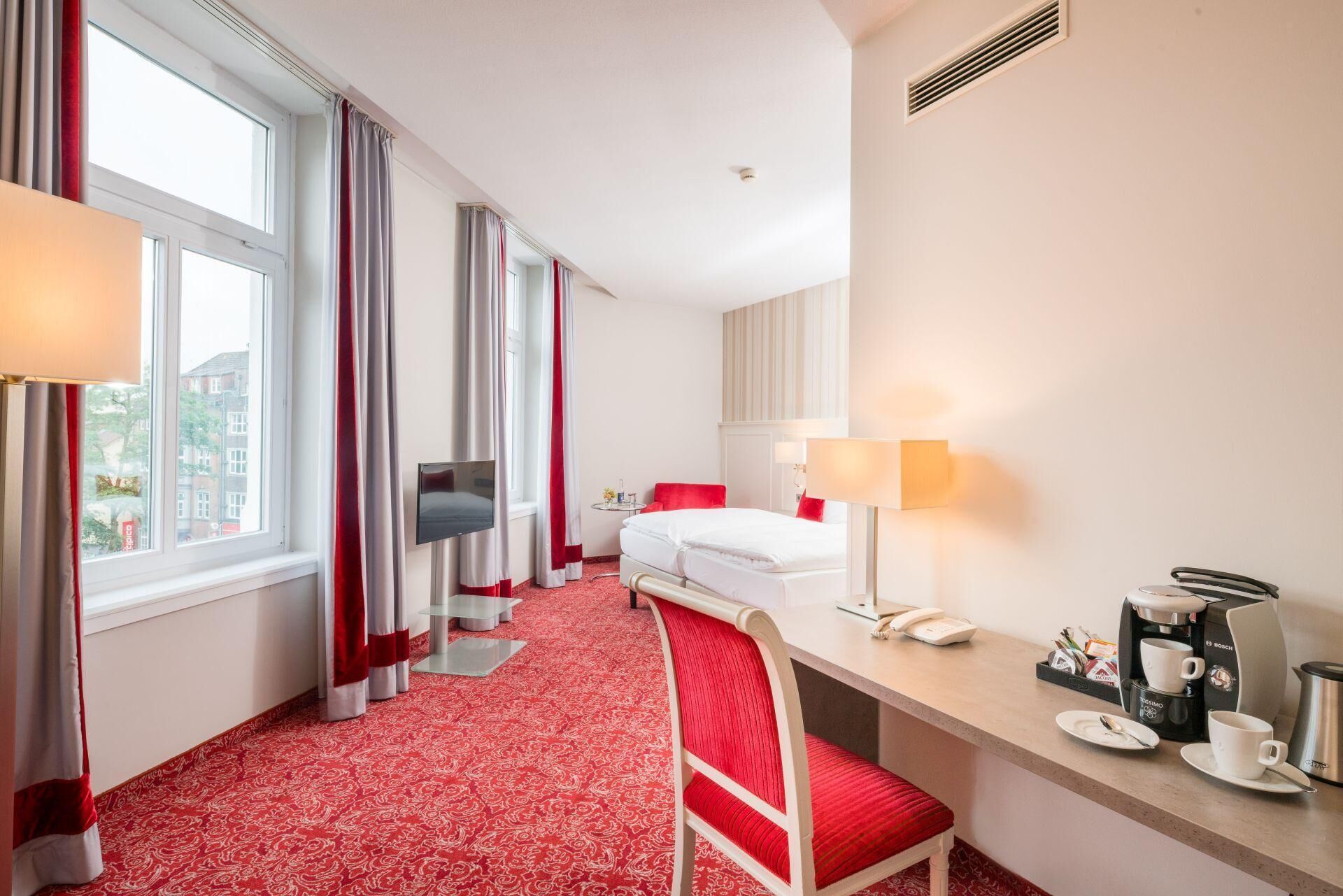 Habitación de hotel con cama, escritorio con teléfono y cafetera, una silla y televisor montado en la pared.