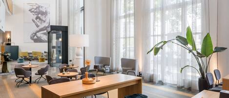 A modern hotel lobby with a central wooden table, bar stools, and a large plant.