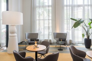 Een moderne hotellobby met een ronde tafel, twee fauteuils, een vloerlamp en een grote kamerplant.