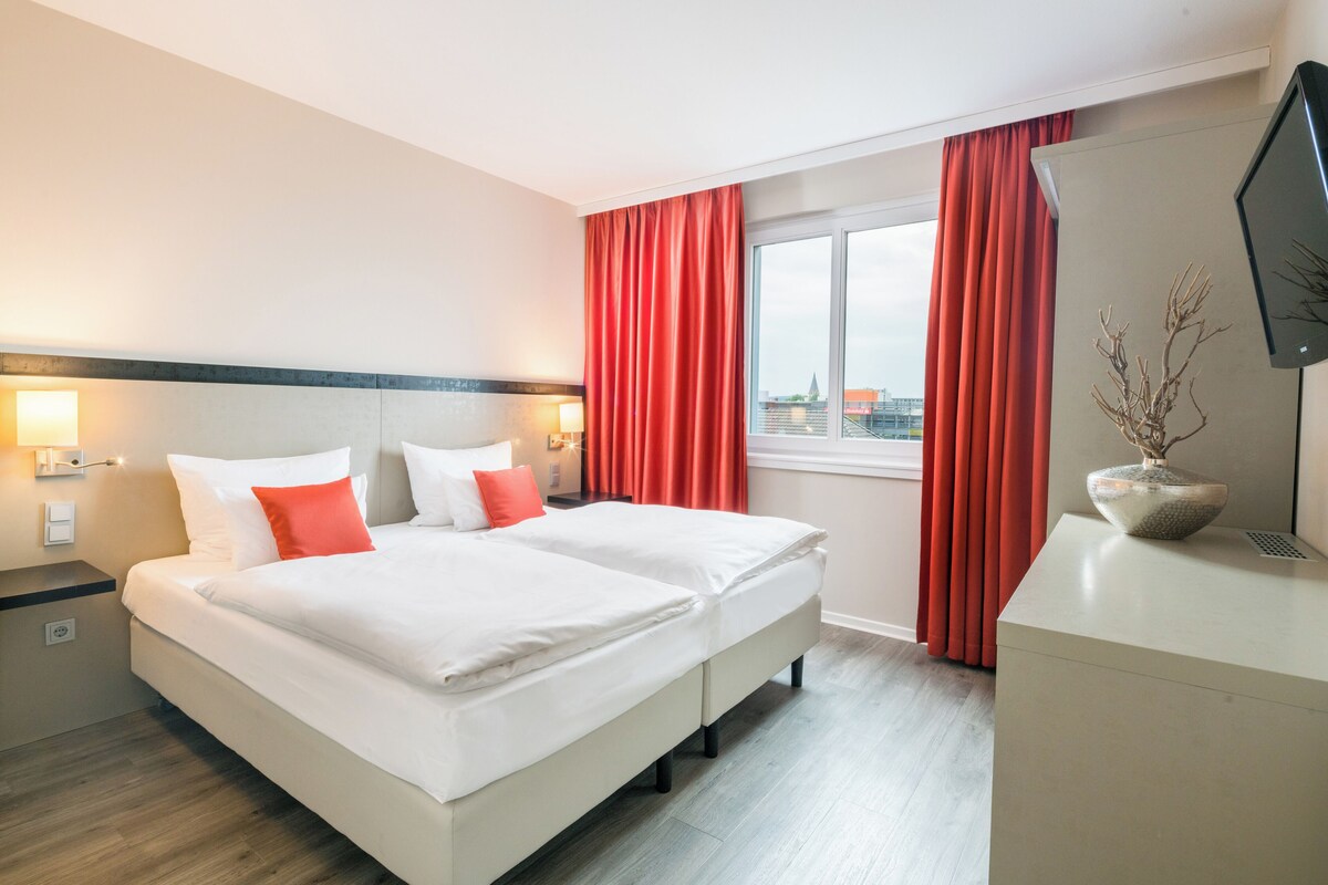 Habitación de hotel con cama, mesita de noche, televisión de pantalla plana, ventana con cortinas y vista a la ciudad.