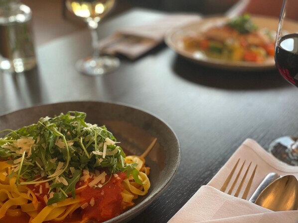 A dining table with a plate of pasta, a glass of red wine, and a napkin with cutlery.