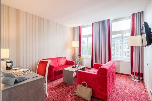 A hotel room with a red sofa set, a small table with a lamp, a desk with a laptop, and a TV mounted on the wall.