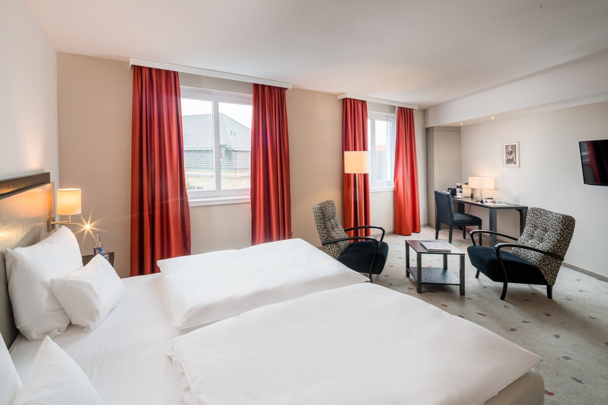 Habitación de hotel con dos camas, un escritorio, dos sillas, un televisor y una ventana con cortinas.
