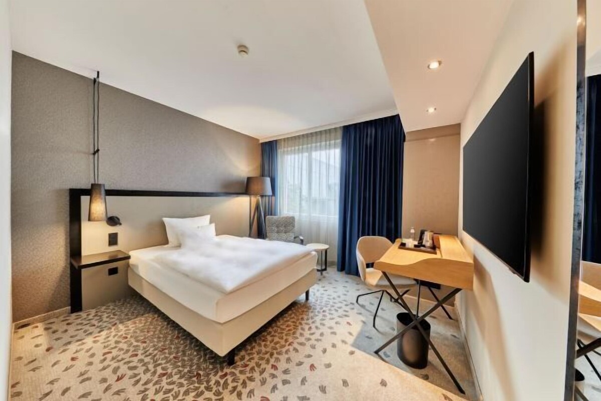 Habitación de hotel con cama, escritorio, silla, televisión y ventana con cortinas.