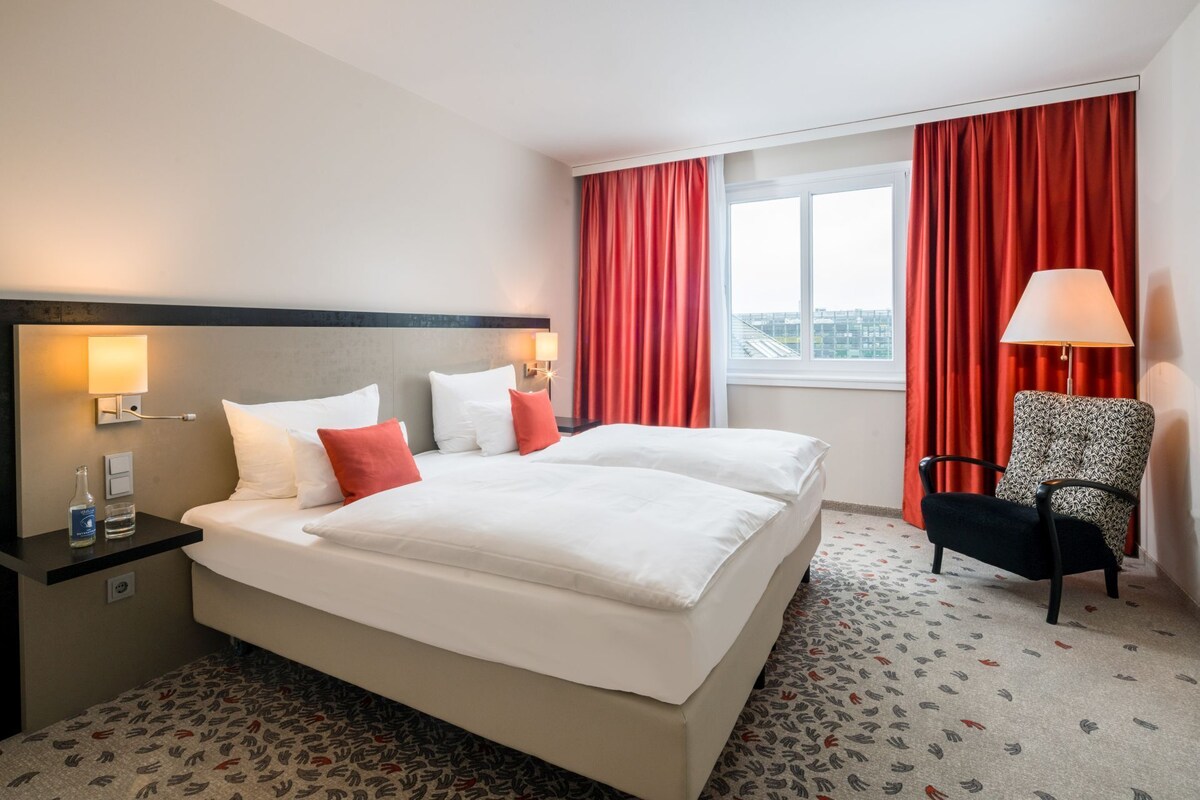 Habitación de hotel con cama doble, mesita de noche con lámpara, una silla y ventana con cortinas rojas.