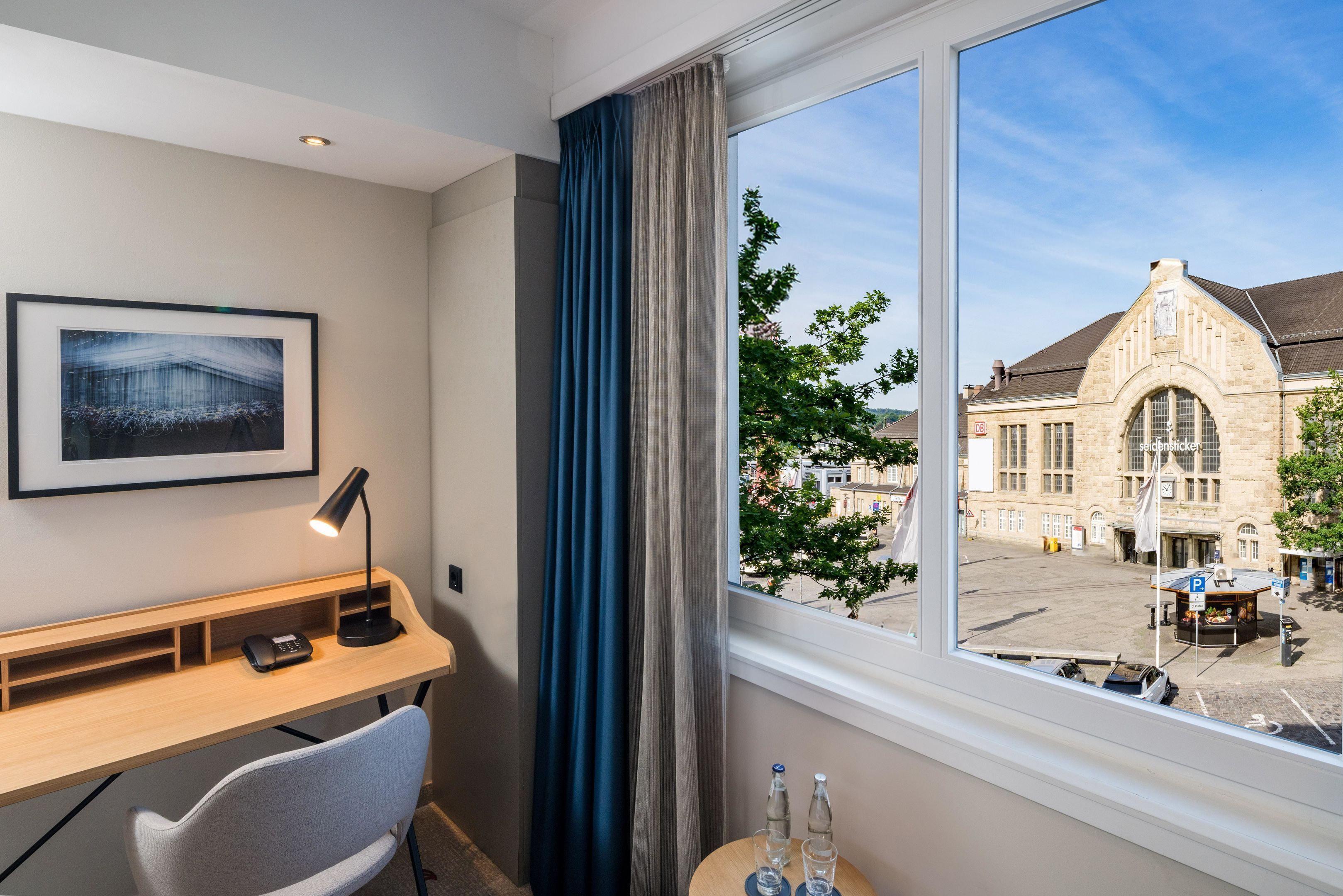 Habitación de hotel con escritorio, silla, un cuadro en la pared, ventana con vistas a un edificio y árboles, y una mesita con dos botellas y vasos.