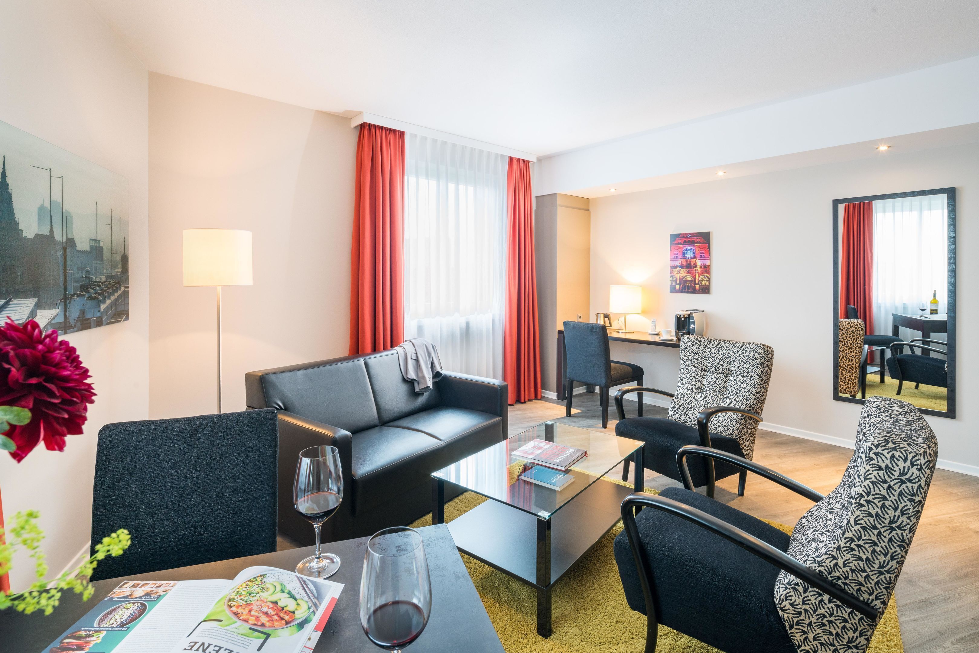 Ein modernes Hotelzimmer mit Sofa, Sessel und Esstisch, auf dem sich eine Zeitschrift und ein Weinglas befinden.