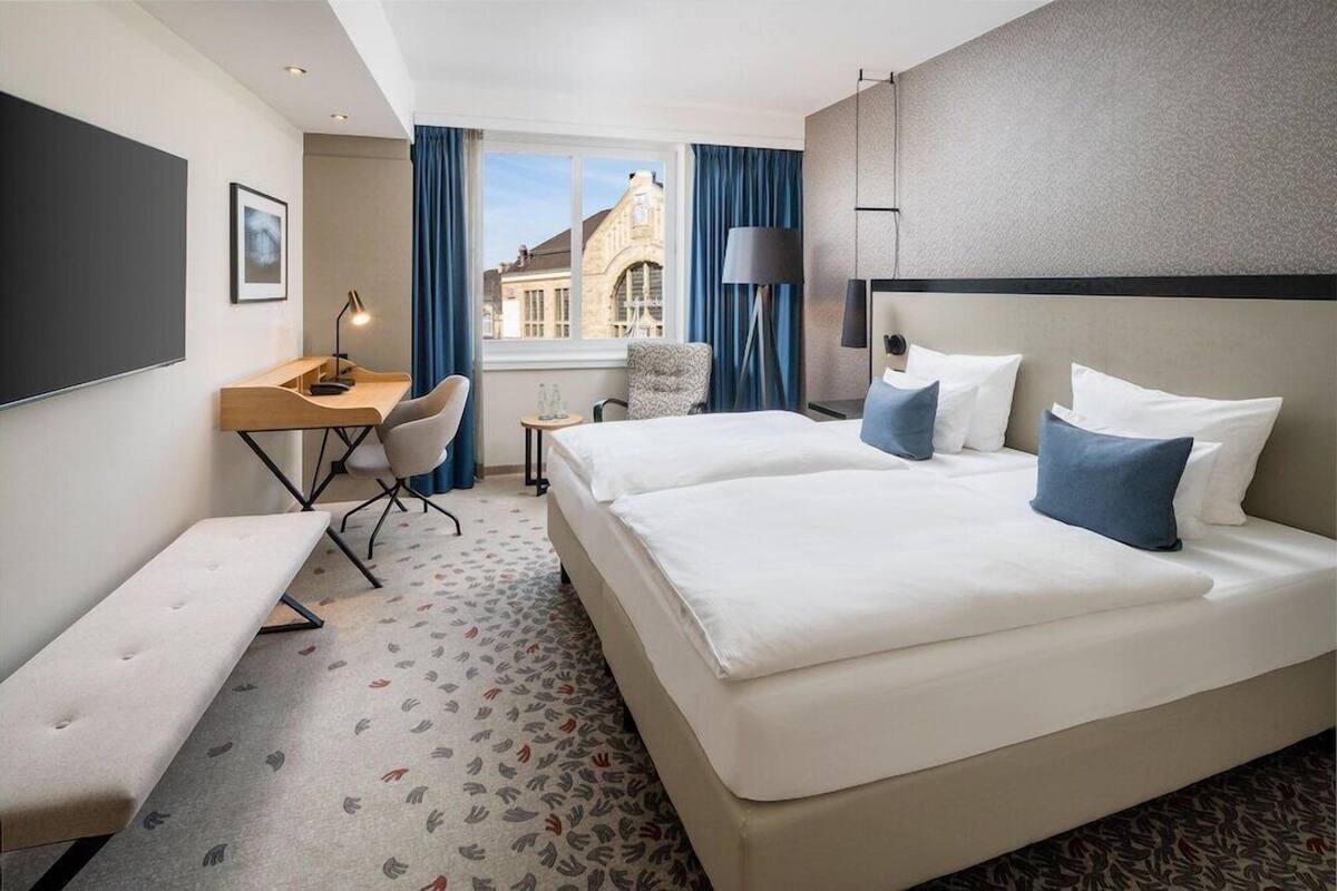 Een hotelkamer met een groot bed, een bureau, een stoel, een bankje en uitzicht op een gebouw.