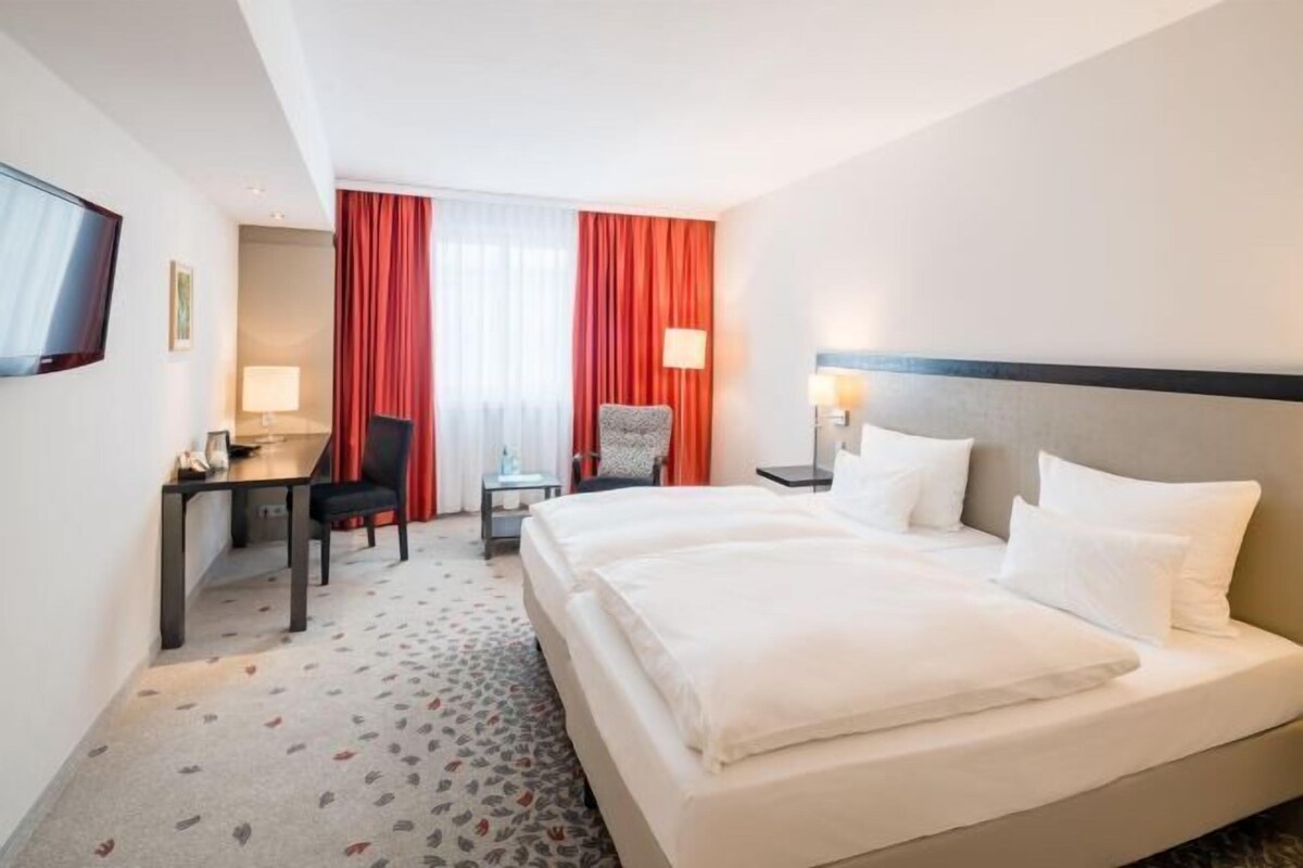Habitación de hotel con cama, escritorio, silla, televisor y ventana con cortinas.