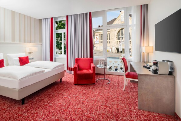 Habitación de hotel con una cama grande, un sillón rojo, un escritorio con una lámpara y vista a un edificio a través de la ventana.
