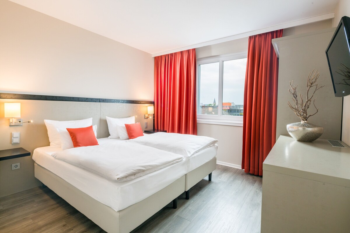 Una habitación de hotel con cama, escritorio, ventana con cortinas y vista a la ciudad.