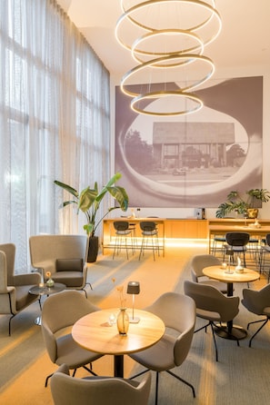 Un moderno vestíbulo de hotel con mesas redondas, asientos cómodos, una planta grande y una lámpara de techo original.