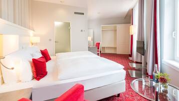 Habitación de hotel con una cama grande, zona de estar con una silla roja, una mesa de vidrio y televisión.