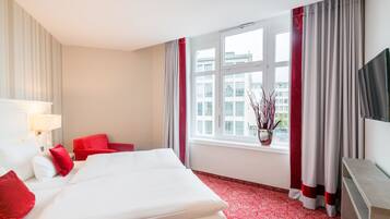 A hotel room with a bed, red pillows, a TV, a window with curtains, and a vase with branches.