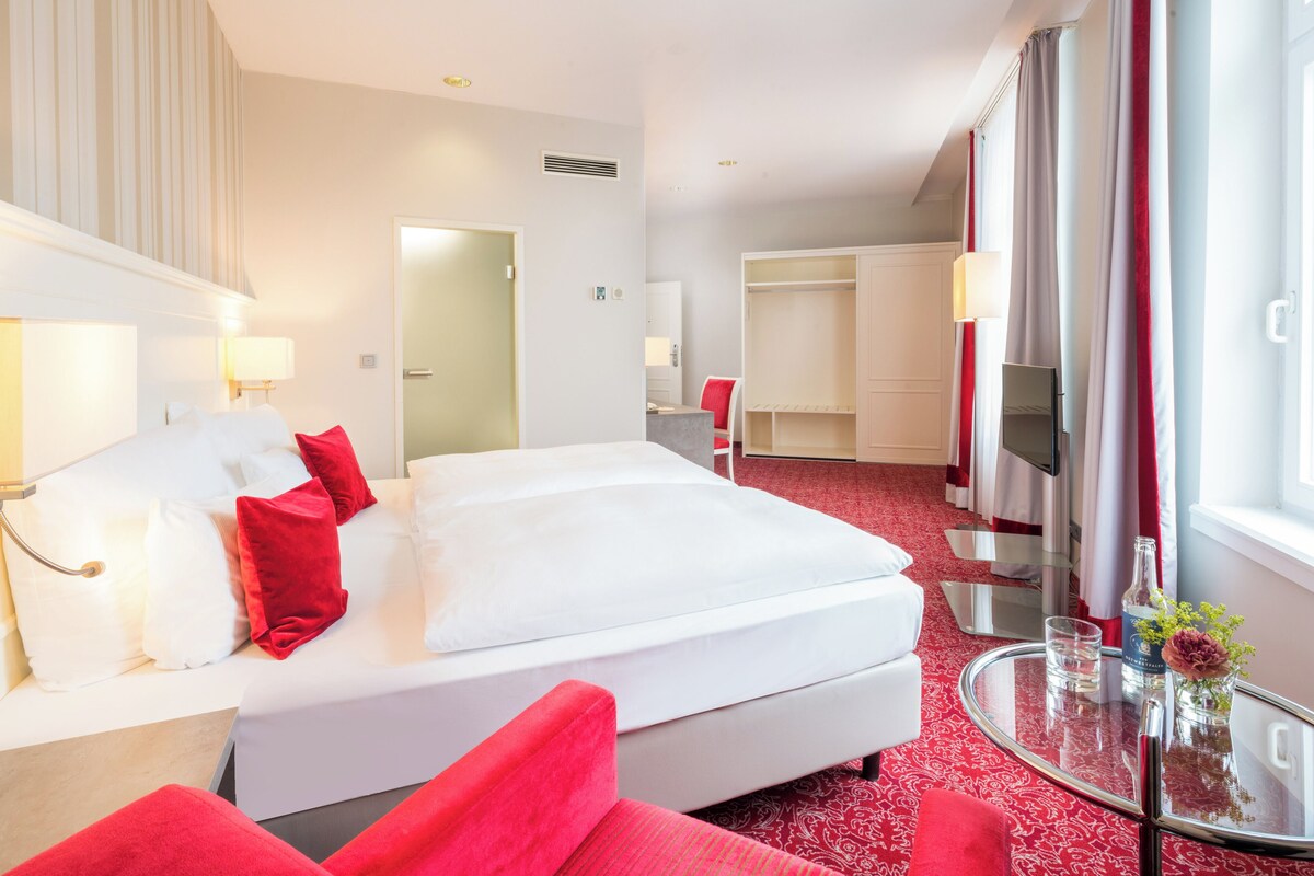 Una habitación de hotel con una cama grande, un sillón rojo, una mesa de centro de vidrio y un televisor.