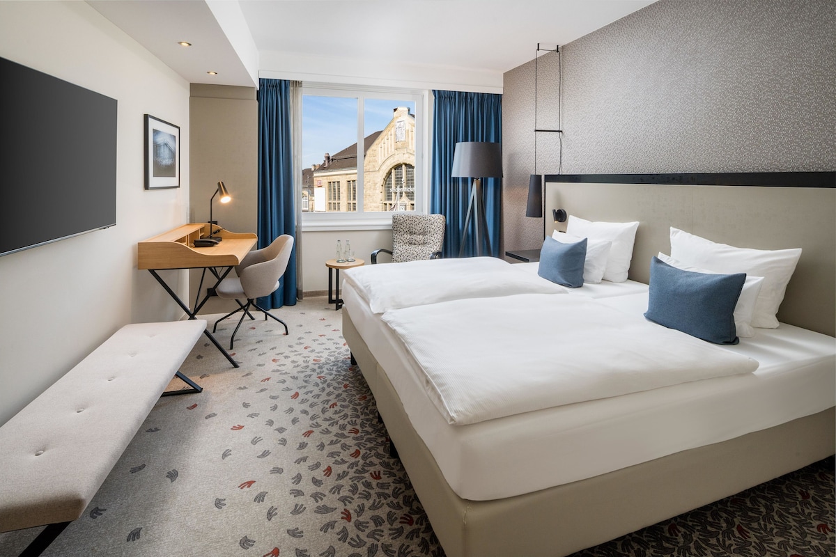 Habitación de hotel con una cama grande, un escritorio, una silla, un banco y vistas a un edificio desde la ventana.
