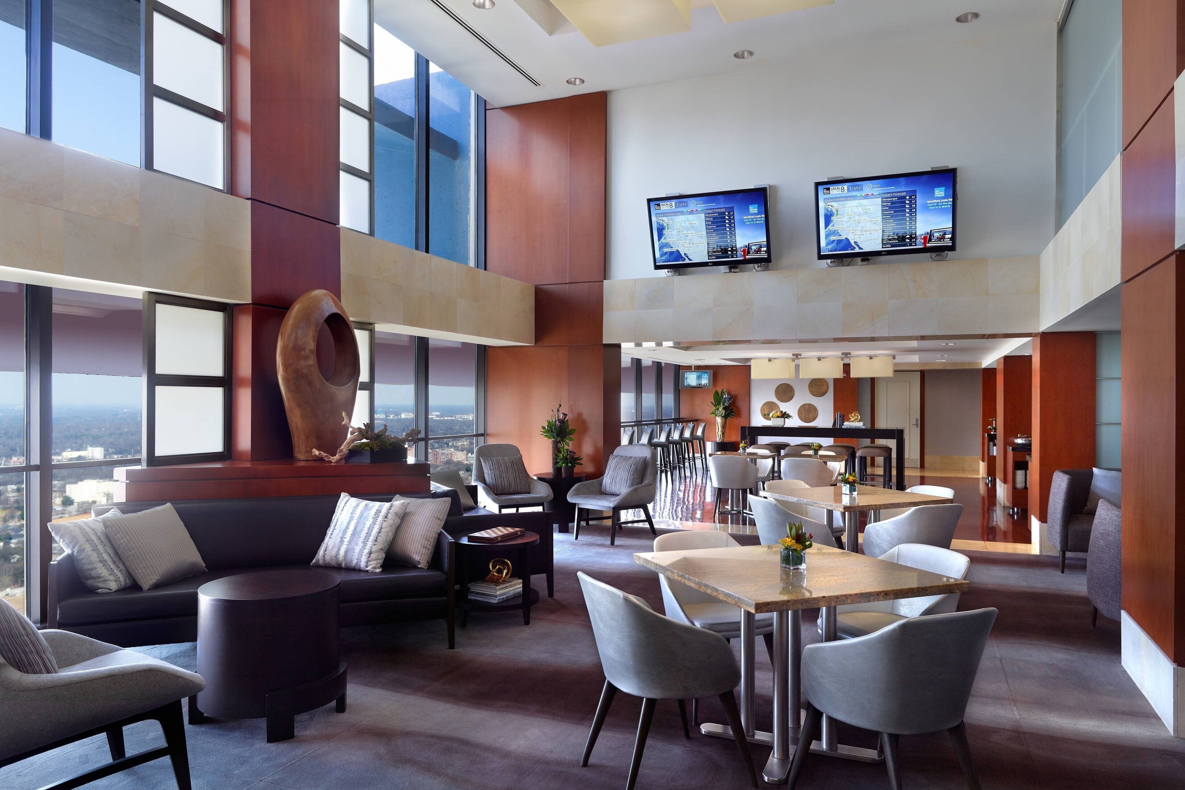 A modern hotel lobby with a high ceiling, large windows, a seating area with sofas and chairs, a dining area with tables and chairs, and two mounted televisions.