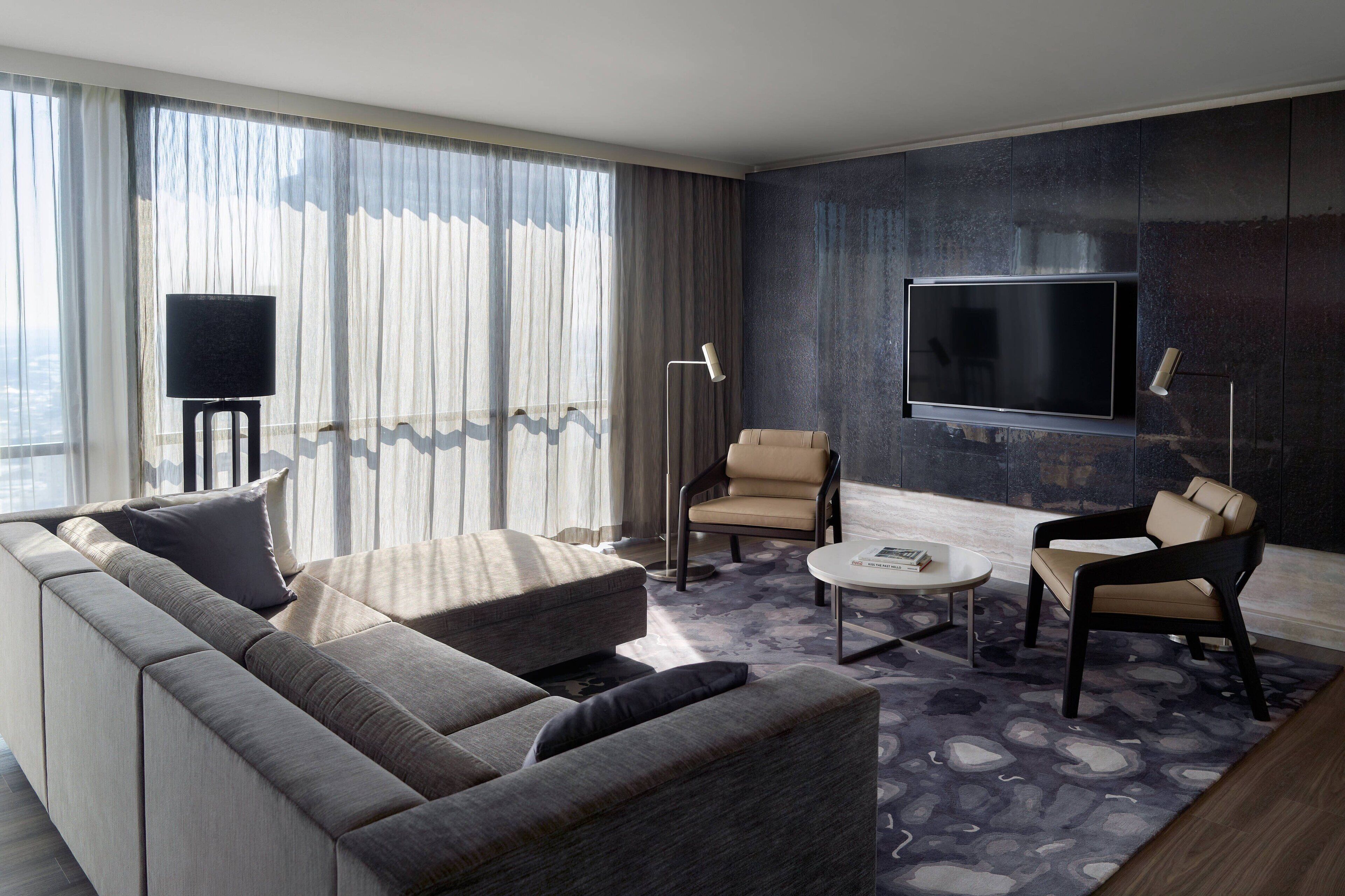 A modern living room with a sofa, two armchairs, a coffee table, a flat-screen TV mounted on the wall, and a floor lamp.