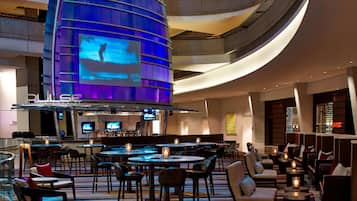 Eine moderne Hotelhalle mit gewölbter Decke, verschiedenen Sitzbereichen und einer Bar mit großem Bildschirm, der Inhalte zeigt.