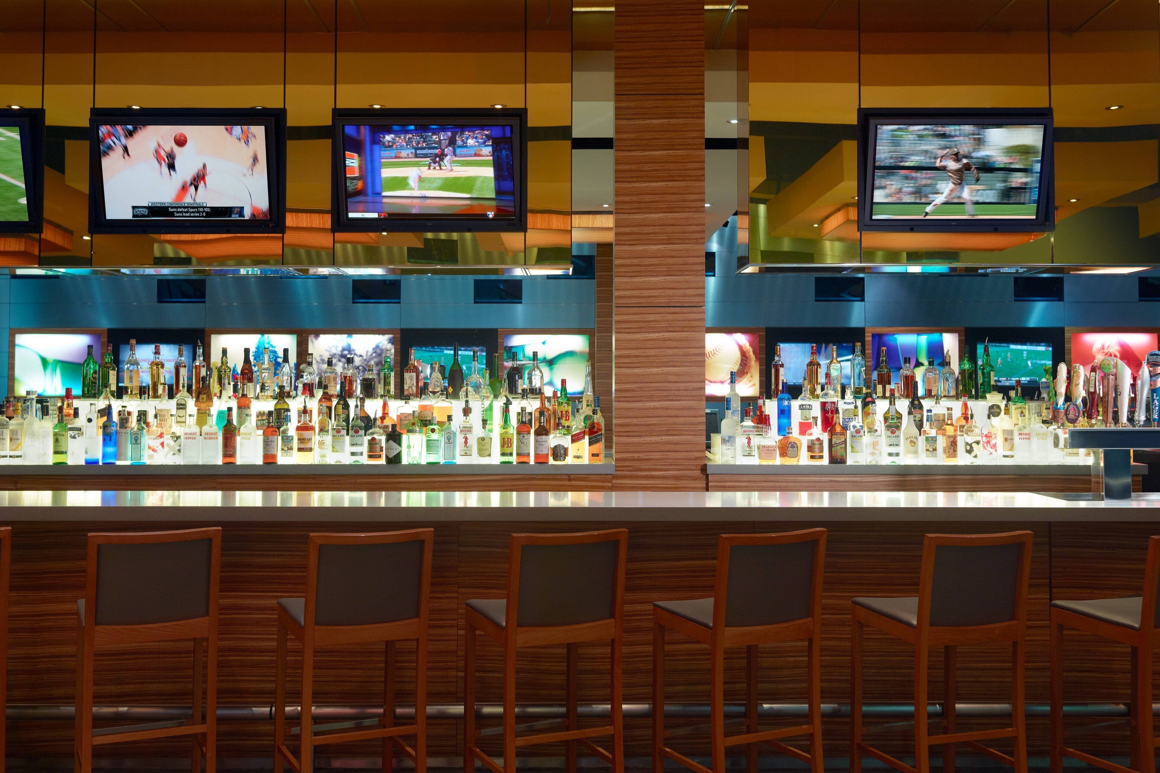 A bar area with multiple screens displaying sports events, a variety of liquor bottles, and wooden bar stools.