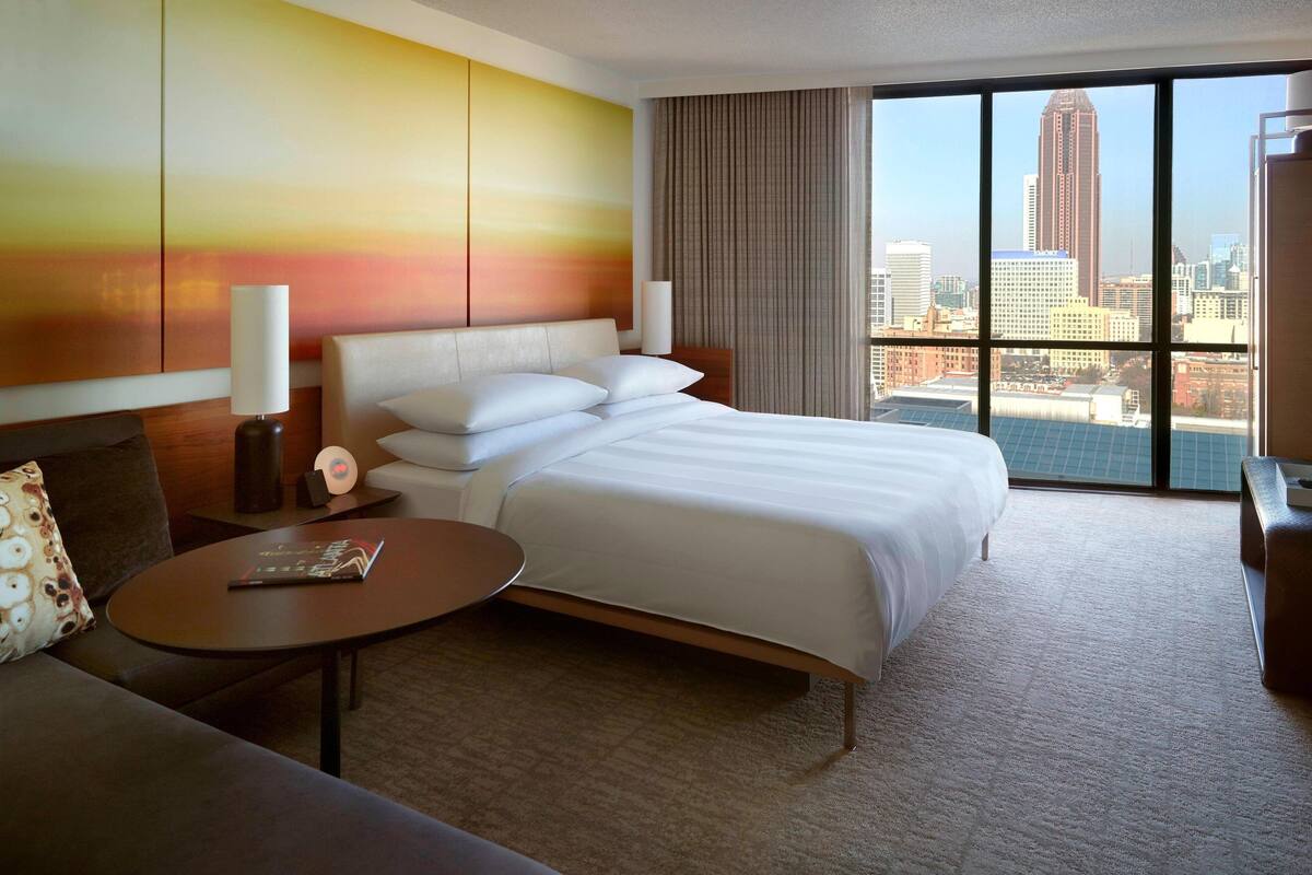 Een hotelkamer met een groot bed, een slaapbank, een ronde tafel en uitzicht op de stad door grote ramen.