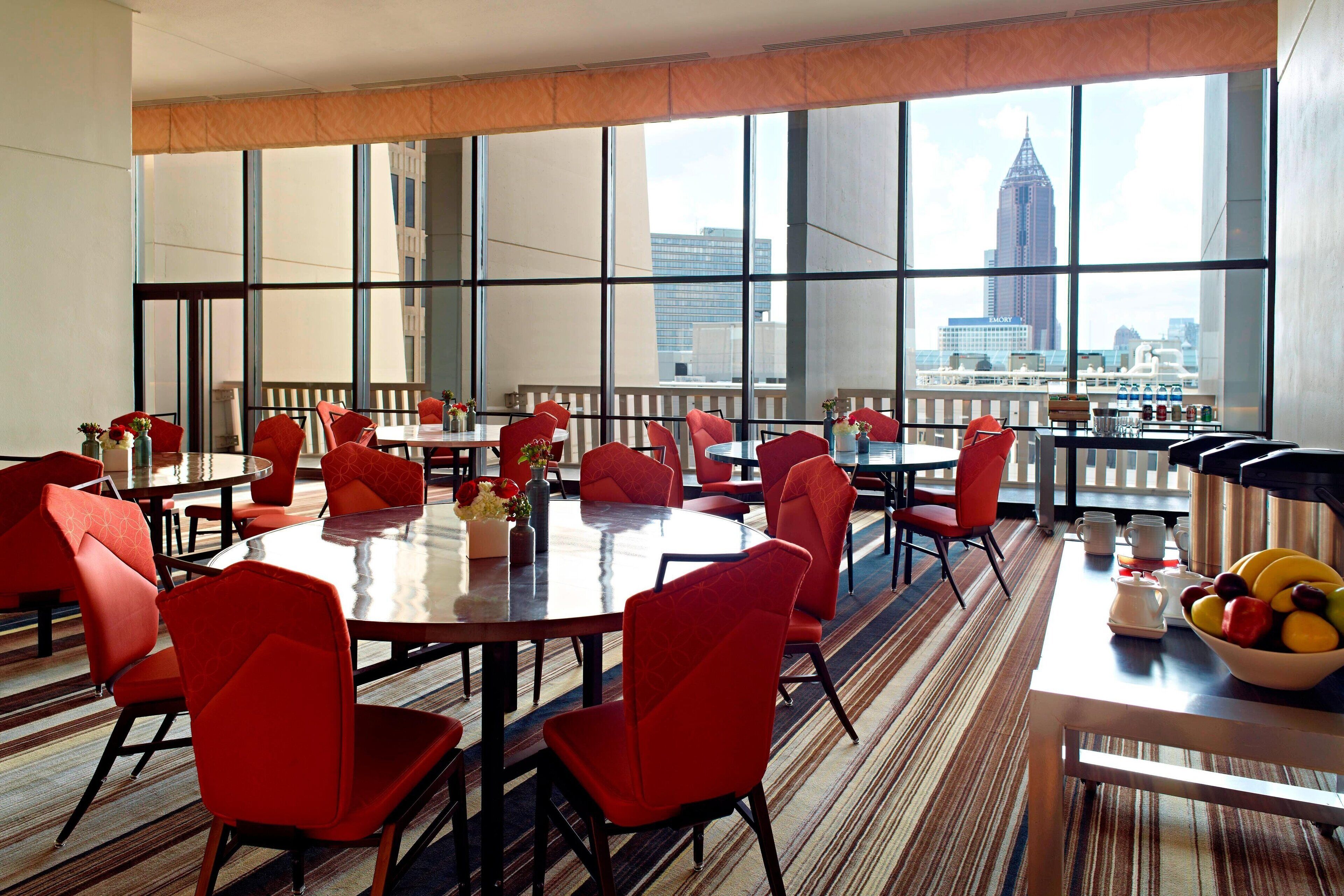 A hotel dining area with multiple tables and red chairs, large windows with a city view, and a bowl of fruit on one table.
