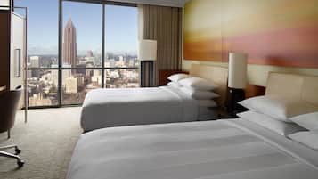 A hotel room with two beds, a desk, a chair, and a large window offering a city view.