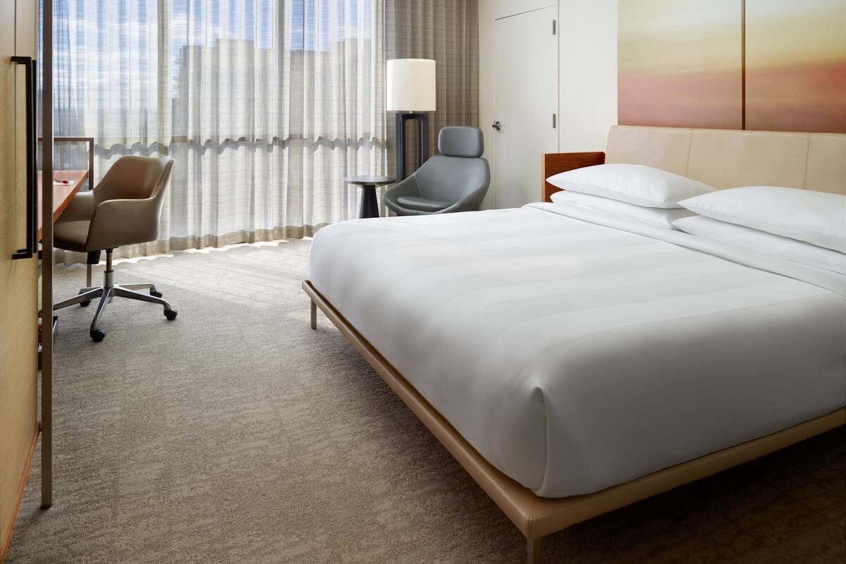 Een hotelkamer met een groot bed, een bureau met een stoel, een lamp en een uitzicht op de stad door dunne gordijnen.
