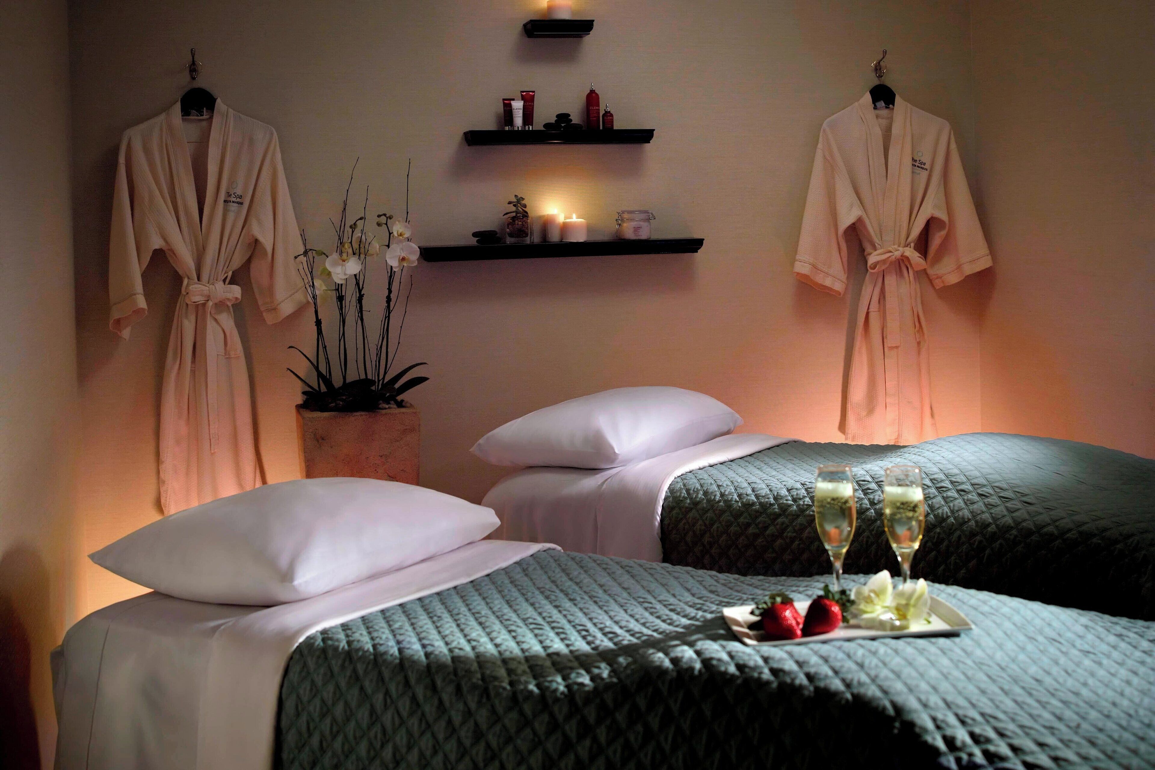 A spa room with two beds, a shelf with candles and toiletries, and two robes hanging on the wall.