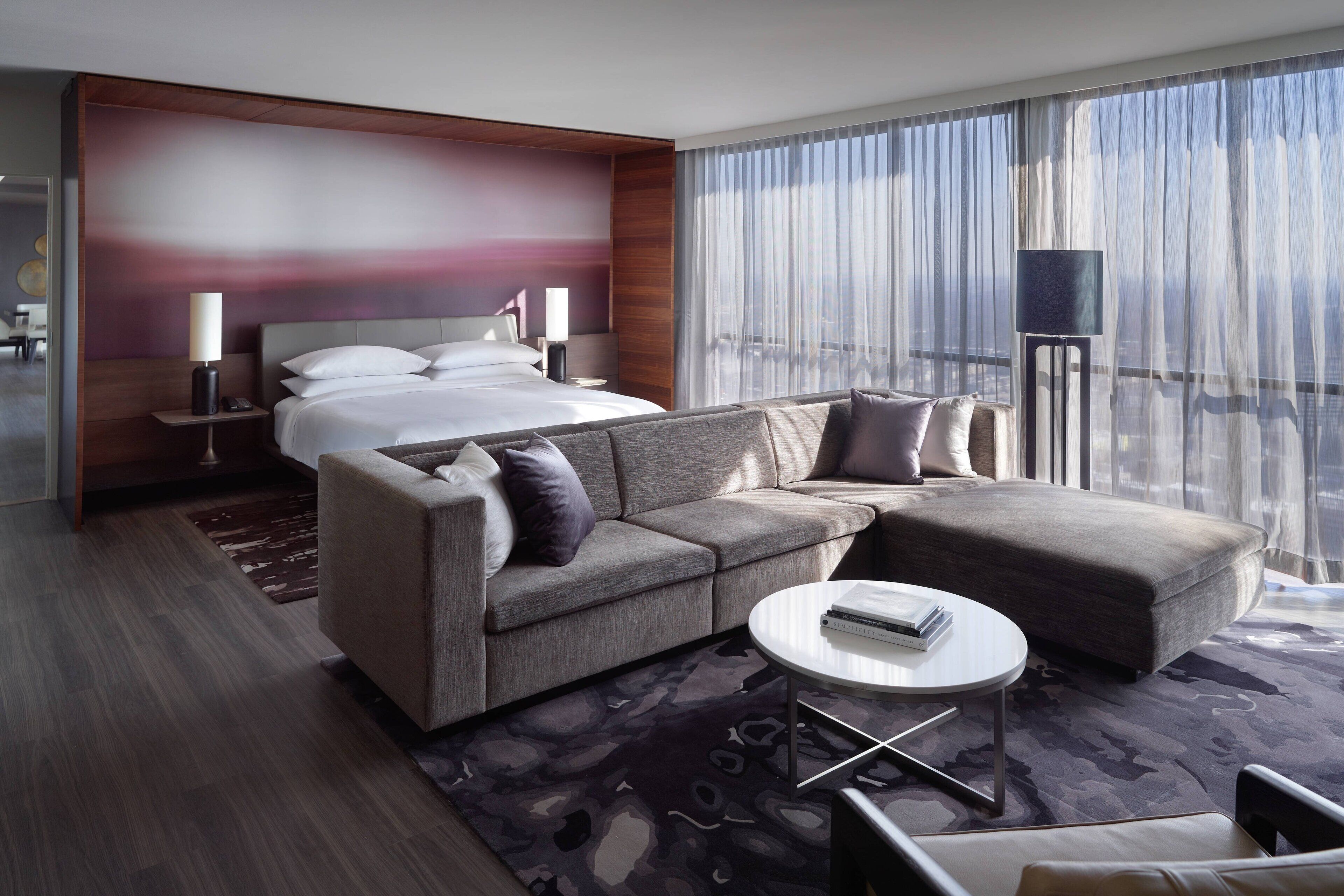 A modern hotel room with a large bed, a sofa, a coffee table, and a view of the city through large windows.