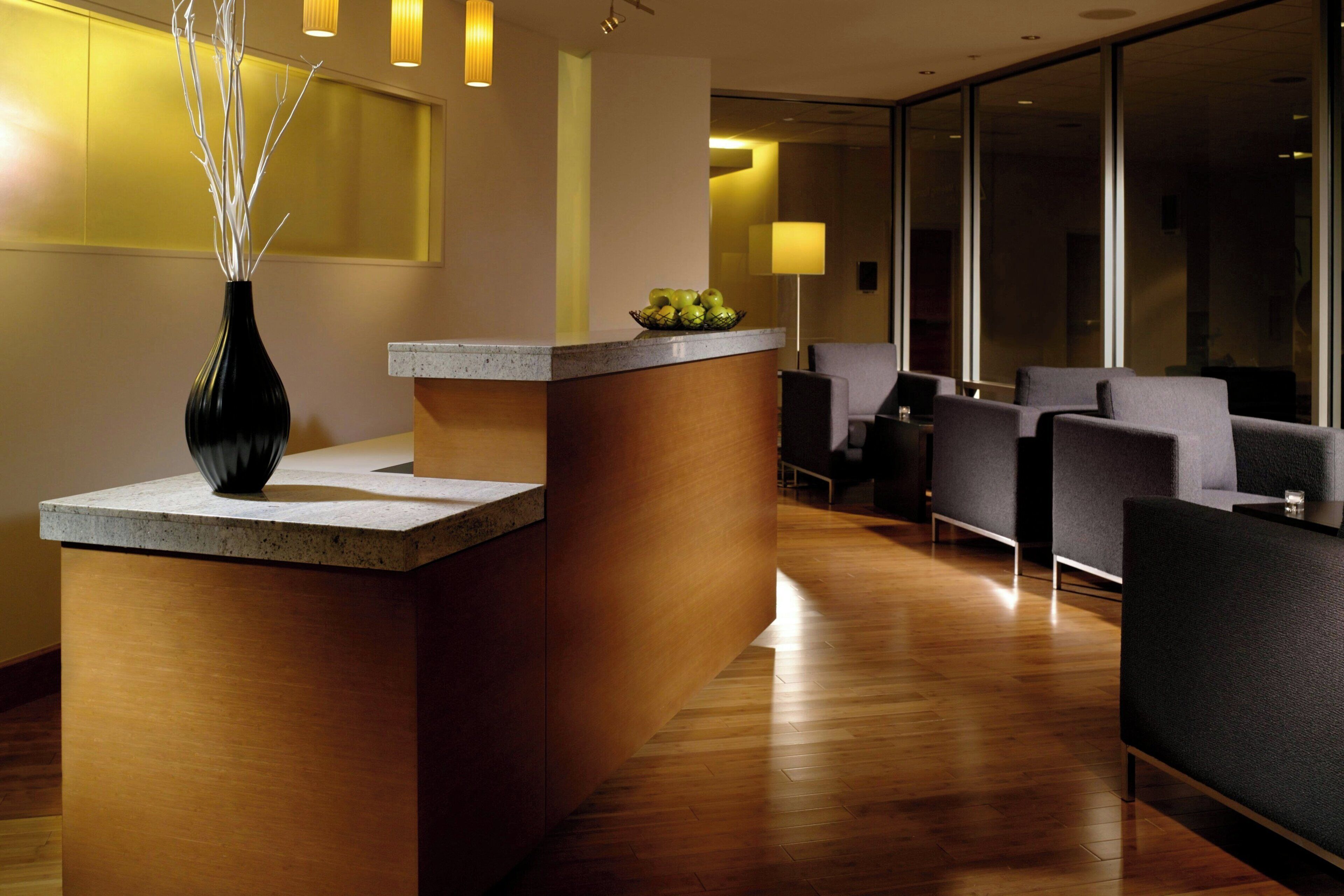 A reception area with a wooden reception desk, a vase with branches, and a bowl of fruit. There are grey chairs and a large window in the background.