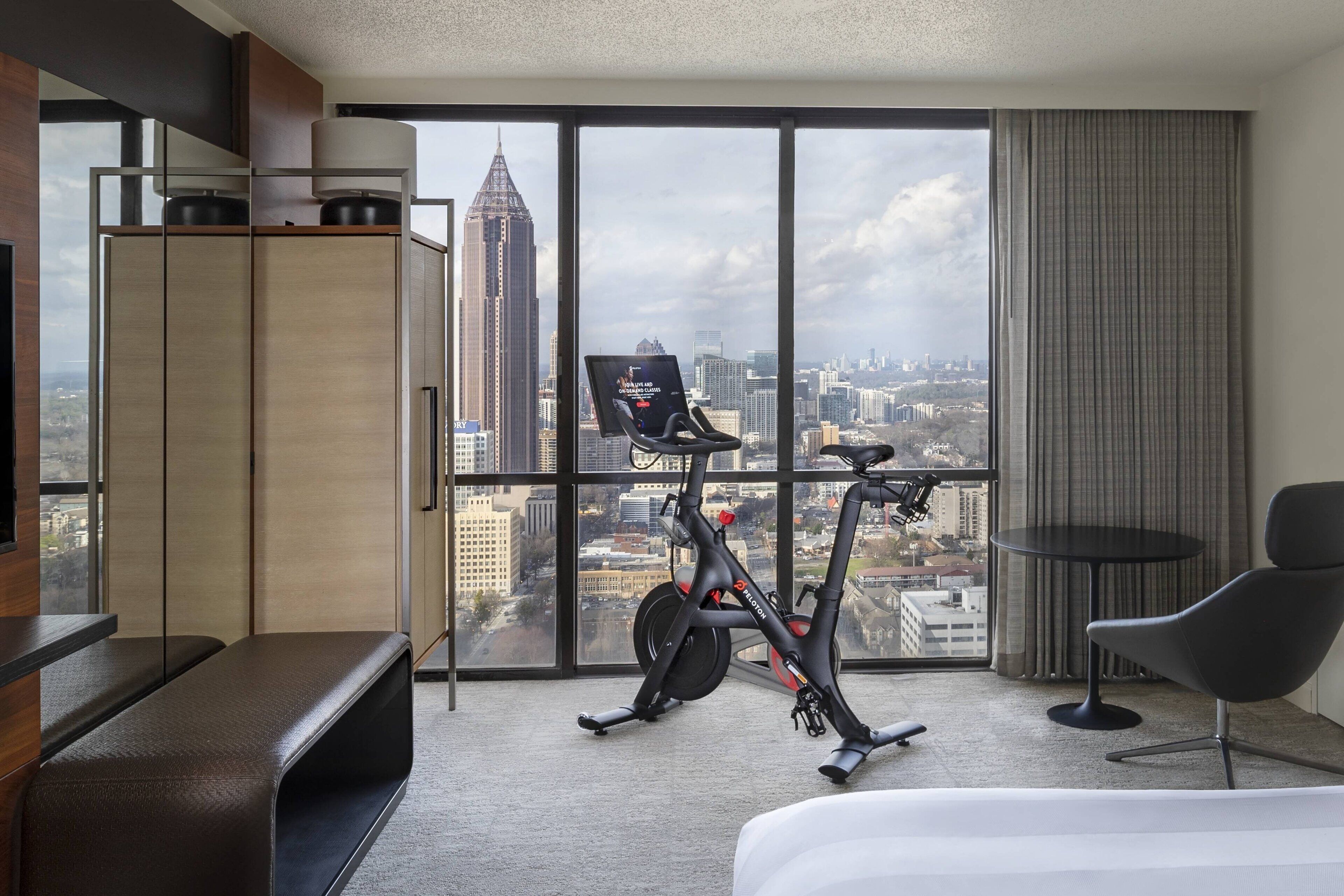 A hotel room with a large window, a treadmill, a bench, a chair, and a bed.