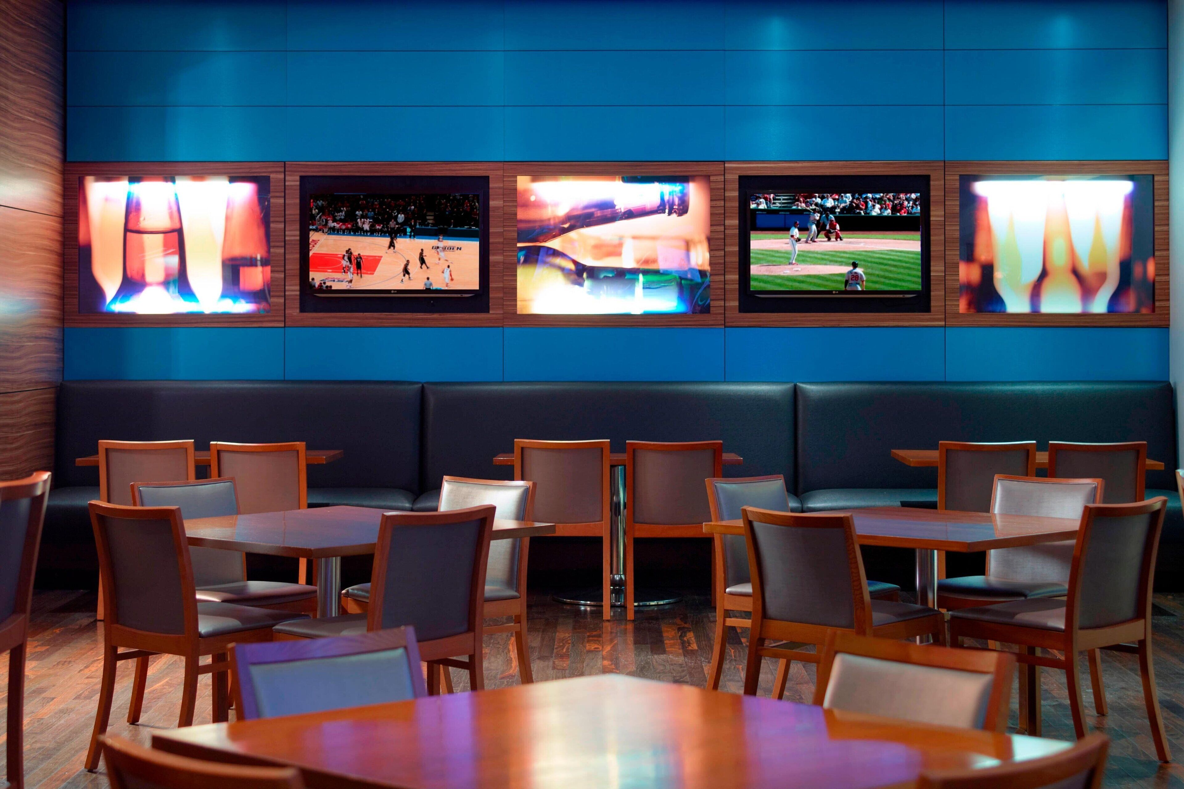 Ein Restaurant mit mehreren Bildschirmen, die Sportereignisse zeigen, Holztischen und -stühlen sowie einer dunkelblauen Wand.