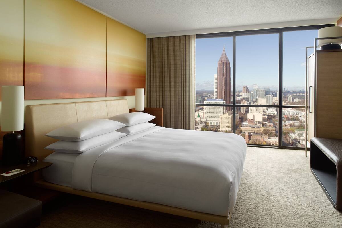 Een hotelkamer met een groot bed, een nachtkastje, een bureau en uitzicht op een stadsgezicht door grote ramen.