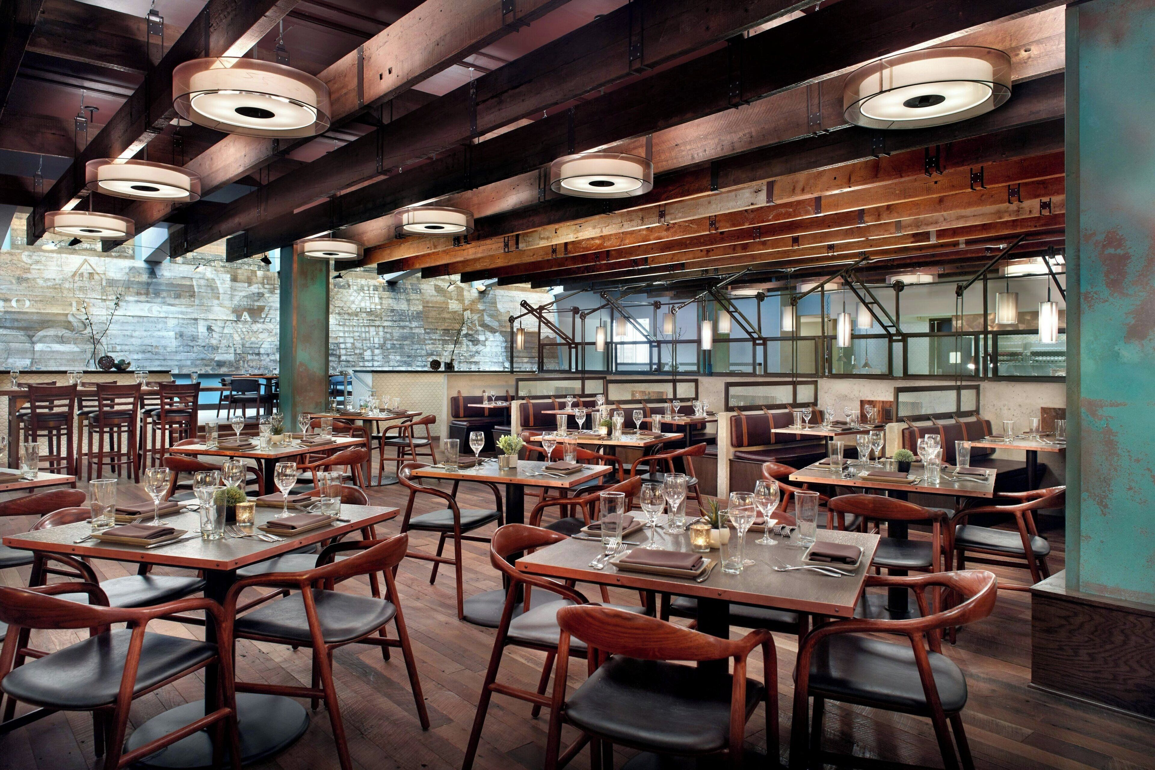 A spacious restaurant with high ceilings, exposed beams, and large windows. Tables are set with wine glasses and menus.