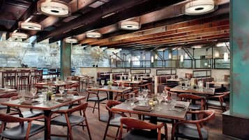 Um restaurante espaçoso com pé-direito alto, vigas aparentes e grandes janelas. As mesas estão dispostas com copos de vinho e cardápios.