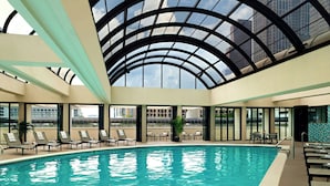 Una piscina interior con sillas reclinables y mesas, ventanas amplias y un techo de cristal.