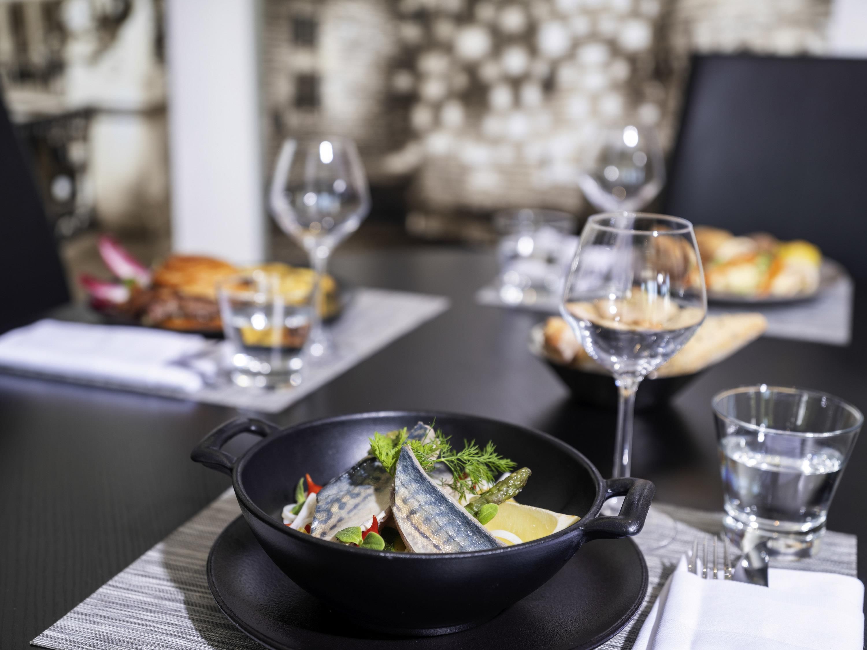 A dining table with a black skillet containing fish, garnished with herbs and lemon, and a glass of water.