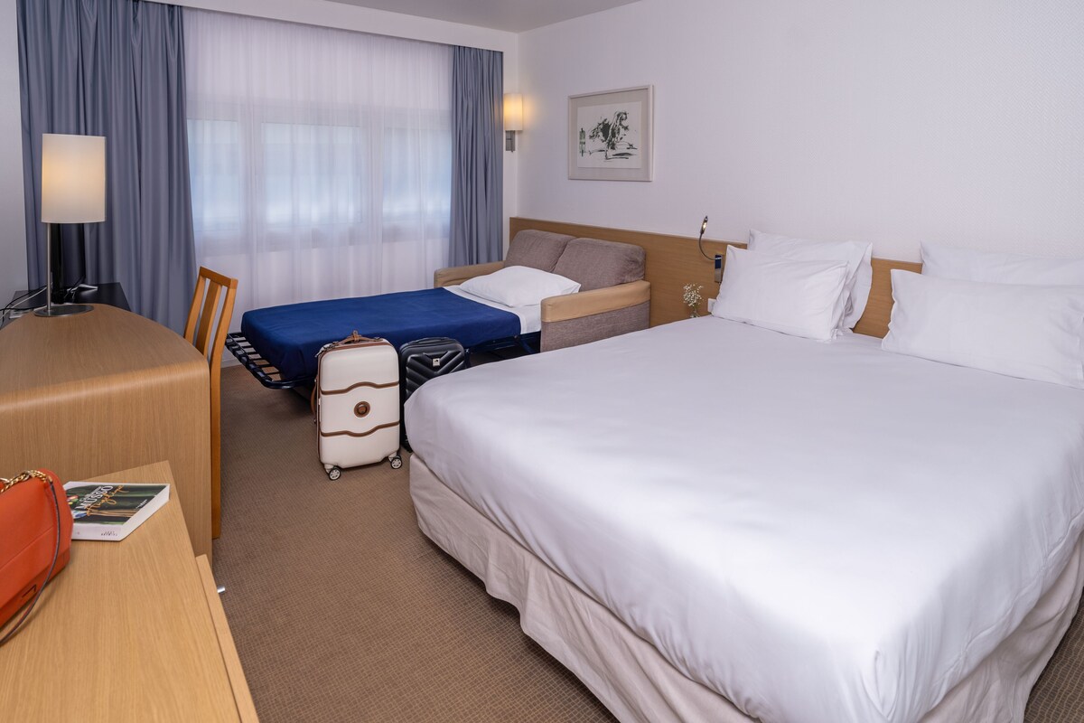 Een hotelkamer met twee bedden, een bureau en een stoel. Er is een raam met gordijnen, een lamp en een lijst met een afbeelding aan de muur.
