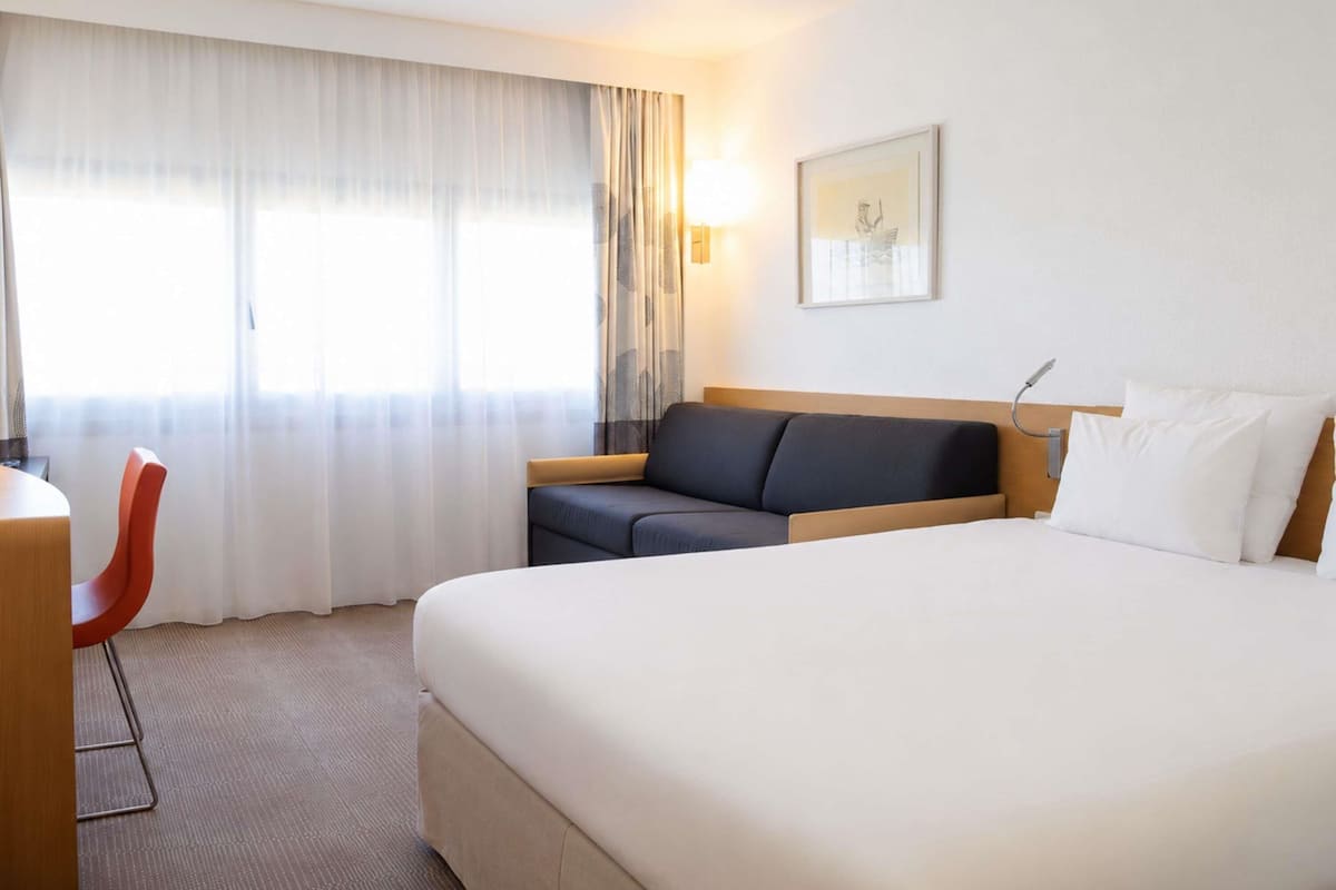 A hotel room with a large bed, a desk, a chair, a sofa, and a window with curtains.