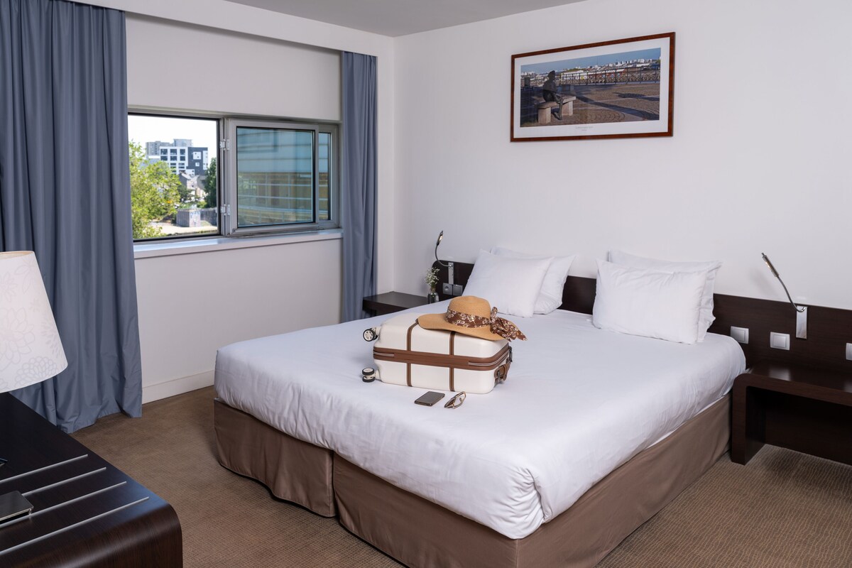Hotelkamer met een groot bed, nachtkastje, bureau, lamp, raam met gordijnen en een lijst met een afbeelding aan de muur.