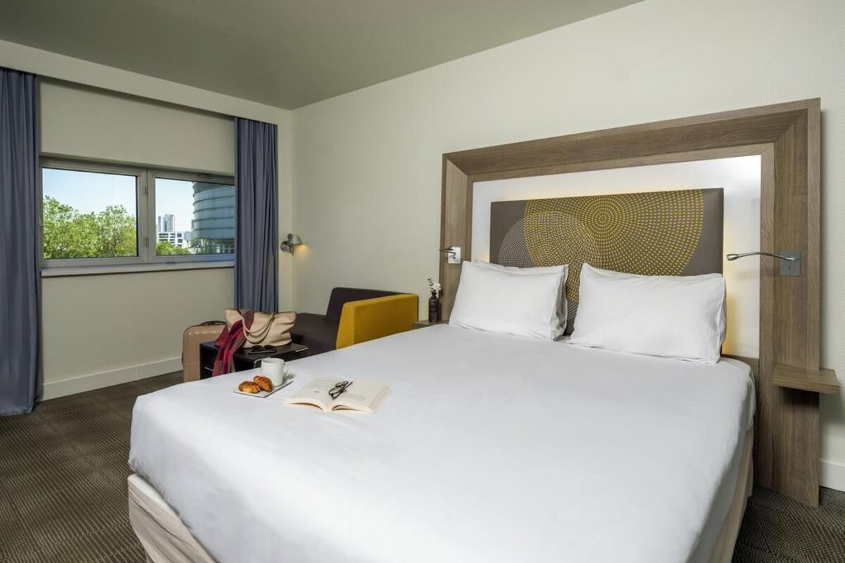Een hotelkamer met een groot bed, een bureau met stoel, een raam met gordijnen en een wandlamp.